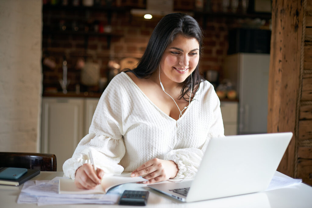 Cheerful young woman teacher with curvy body and black hair working from home having online lesson using earphones and laptop webcam, handwriting in notebook, smiling. Distant education concept